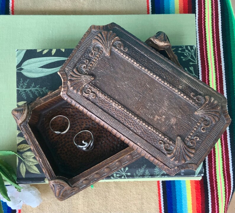 May include: A vintage wooden ring box with an ornate design. The box is open and has two silver rings inside.