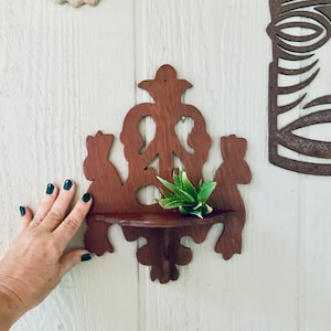 May include: A dark brown wooden shelf with intricate carvings in the shape of leaves and flowers. The shelf has a small ledge for displaying items and is mounted on a white wall. A small green succulent plant is sitting on the shelf.