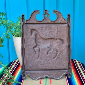May include: A brown metal wall plaque with a raised design of a horse. The plaque has a decorative frame with a scalloped edge.