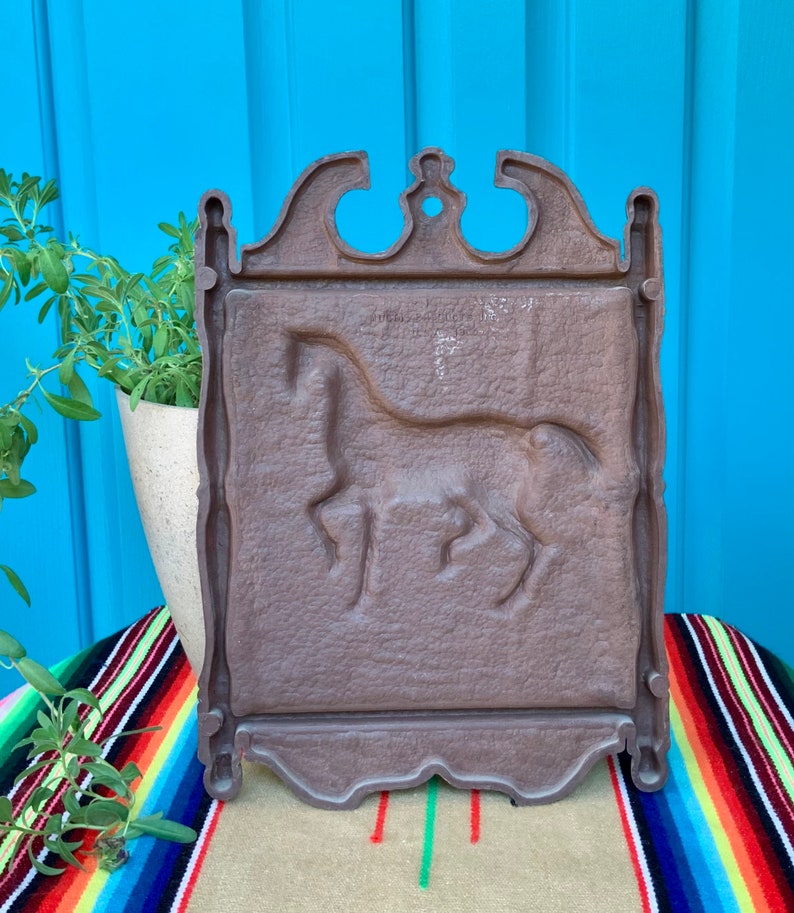 May include: A brown metal wall plaque with a raised design of a horse. The plaque has a decorative frame with a scalloped edge.