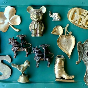 Op de afbeelding: Een verzameling vintage messing snuisterijen op een groene dienblad. De items omvatten een klavertjevier, een Mickey Mouse-figuur, een kleine olifant, een blaadjesvormige schaal, een kandelaar, een laars, een letter 'S', een vogelbeeldje en een decoratief stuk met vleugels.