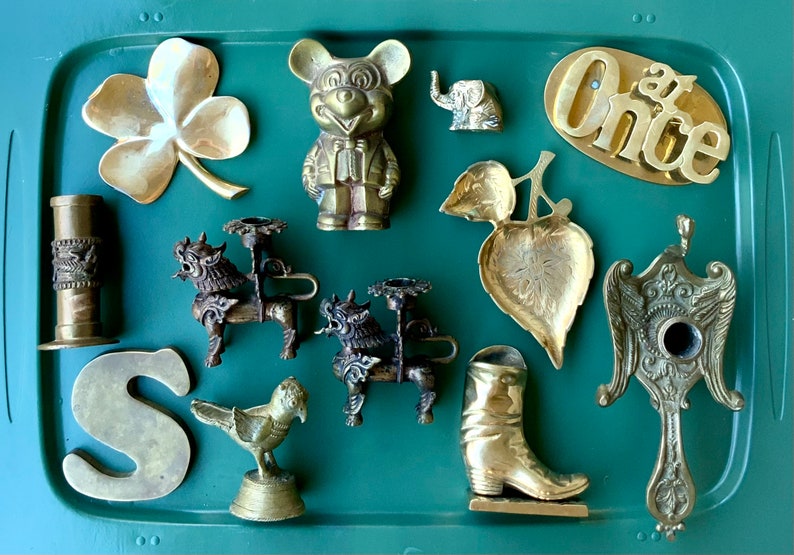 Op de afbeelding: Een verzameling vintage messing snuisterijen op een groene dienblad. De items omvatten een klavertjevier, een Mickey Mouse-figuur, een kleine olifant, een blaadjesvormige schaal, een kandelaar, een laars, een letter 'S', een vogelbeeldje en een decoratief stuk met vleugels.