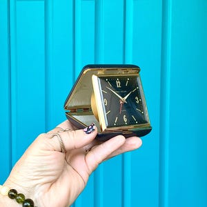May include: A vintage travel alarm clock with a black face, gold accents, and a black leatherette case. The clock face has white numerals and hands, with a red second hand. The clock is open, revealing the gold-colored clock mechanism.