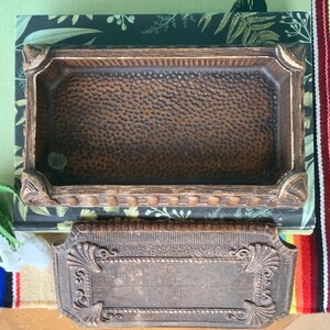 May include: A vintage wooden box with a textured lid and a brown interior. The box has a decorative design around the edges and a raised, ornate design on the lid.