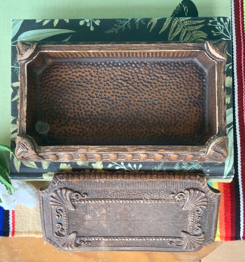 May include: A vintage wooden box with a textured lid and a brown interior. The box has a decorative design around the edges and a raised, ornate design on the lid.