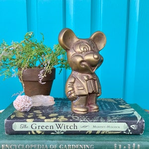 Op de afbeelding: Een messing beeldje van Mickey Mouse met een pak en strikje. Het beeldje staat op een stapel boeken met de titel "The Green Witch" zichtbaar.
