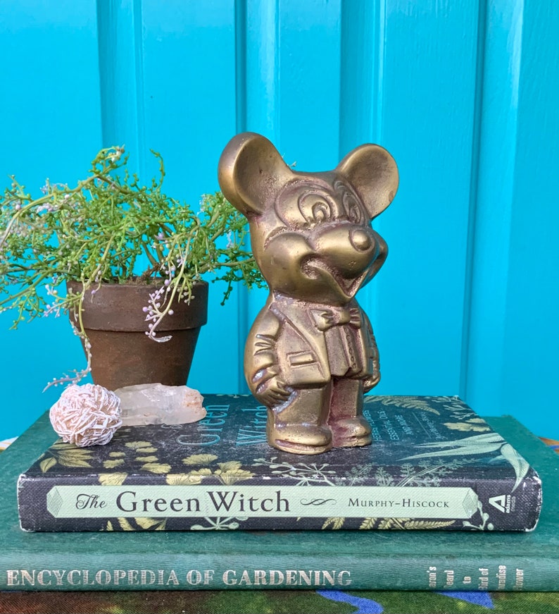 Op de afbeelding: Een messing beeldje van Mickey Mouse met een pak en strikje. Het beeldje staat op een stapel boeken met de titel "The Green Witch" zichtbaar.