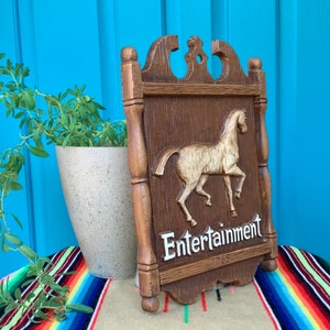 May include: A wooden sign with a carved horse and the words "Entertainment 1765" in white lettering. The sign is brown with a distressed finish and has a decorative top.