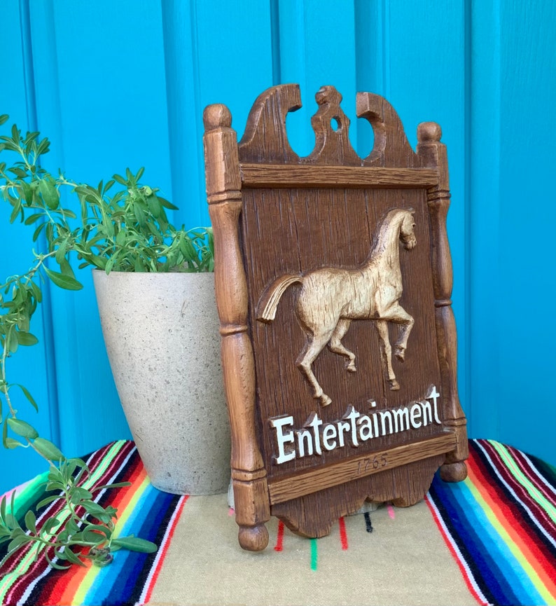 May include: A wooden sign with a carved horse and the words "Entertainment 1765" in white lettering. The sign is brown with a distressed finish and has a decorative top.