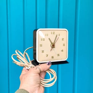 May include: A vintage electric alarm clock with a square face and a black base. The clock has a cream-colored face with gold numerals and hands. The clock's face is framed by a white border. The clock is plugged in with a cream-colored cord.