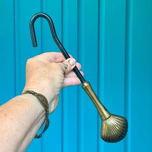 May include: A metal fireplace tool featuring a long, curved black handle and a decorative brass end shaped like a seashell. The tool has a smooth, tapered transition between the black handle and the textured, fan-shaped brass shell component.