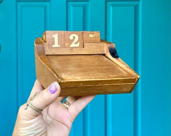 May include: A vintage wooden desk calendar displaying "12 JULY". The calendar is made of light brown wood with a small tray. The numbers and month are printed in a simple, readable font. The calendar is held in front of a teal blue door.