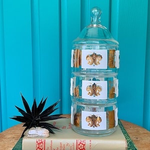May include: A three-tiered clear glass canister with a matching lid, featuring gold-colored decorative floral motifs on each section. The canister rests on a stack of two books, one titled The Teahouse of the August Moon and The North Woods.