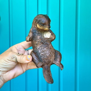 May include: A brown, textured otter figurine depicted in a floating position, holding a small, round, light-colored object against its chest. The sculpture features detailed fur markings and a smooth, rounded tail, capturing the animal in a classic pose.