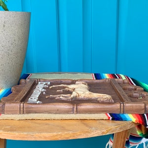 May include: A wooden sign with a carved horse and the text "Estancia" in white lettering. The sign is brown and has a rustic look. It is sitting on a table with a colorful blanket underneath.