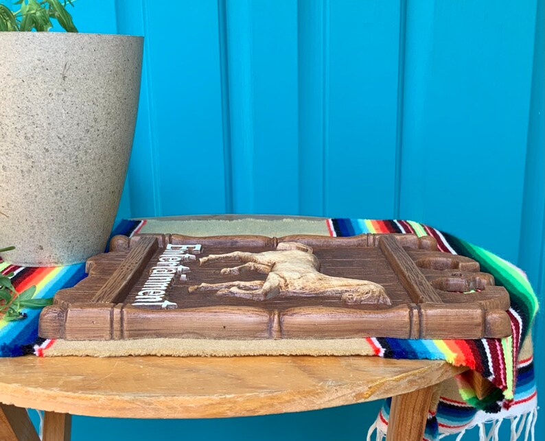 May include: A wooden sign with a carved horse and the text "Estancia" in white lettering. The sign is brown and has a rustic look. It is sitting on a table with a colorful blanket underneath.