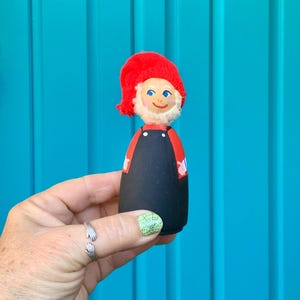 May include: A small wooden gnome figurine with a red knit hat, black overalls, and a painted face. The gnome is holding out its hands.