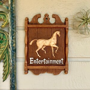 May include: A wooden sign with a carved horse and the words "Entertainment 1765". The sign is brown with a gold horse and white lettering.