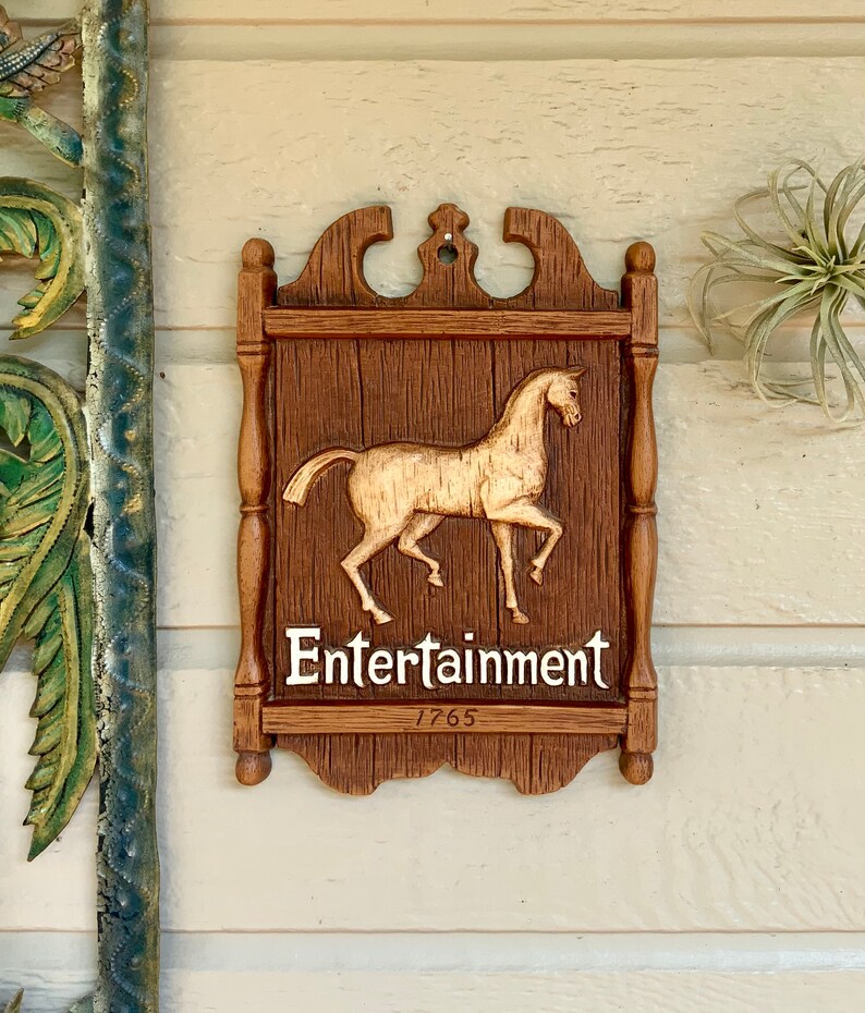 May include: A wooden sign with a carved horse and the words "Entertainment 1765". The sign is brown with a gold horse and white lettering.