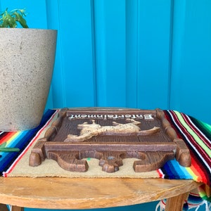 May include: A wooden sign with the word "Entertainment" painted on it. The sign features a carved image of two animals, possibly deer, in a running pose. The sign is brown with a light brown background and is sitting on a colorful striped blanket.
