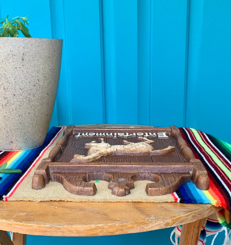 May include: A wooden sign with the word "Entertainment" painted on it. The sign features a carved image of two animals, possibly deer, in a running pose. The sign is brown with a light brown background and is sitting on a colorful striped blanket.