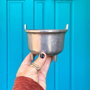 May include: A small, silver, three-legged pot with two handles. The pot is being held in a hand with a gold snake ring.