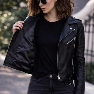 May include: A black leather jacket with a classic biker style, featuring a notched collar, asymmetrical zipper, and silver hardware. The jacket is paired with a black t-shirt and dark wash jeans. The jacket's interior lining is visible.