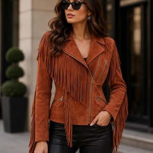 May include: A rust-colored suede jacket featuring long fringe detailing along the chest, shoulders, and sleeves. The jacket includes a notched lapel, an asymmetrical front zipper closure, and two zippered pockets on the front waist area.