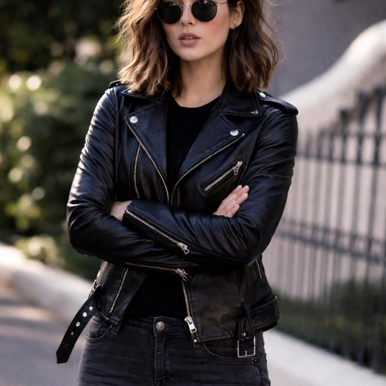 May include: A black leather jacket with silver zippers and a belt. The jacket has a classic biker style with a notched collar and multiple pockets. The person is wearing black sunglasses and dark-wash jeans.