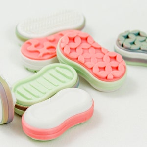 May include: A collection of colorful, decorative erasers in various shapes, including a sneaker sole design, floral patterns, and a simple rectangular shape. The erasers are in shades of pink, green, white, and blue, with a white background.