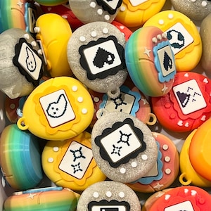 May include: A collection of colorful, retro-style digital pet toys in various colors, including yellow, gray, red, and rainbow. Each toy features a small screen displaying pixelated graphics and a decorative frame. The toys are designed with a rounded, ergonomic shape.