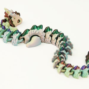 May include: A 3D-printed articulated dragon toy in shades of green, blue, and purple. The dragon has a detailed design with a segmented body, a head with brown and green accents, and a curved tail. The toy is on a white background.