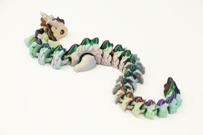 May include: A 3D-printed articulated dragon toy in shades of green, blue, and purple. The dragon has a detailed design with a segmented body, a head with brown and green accents, and a curved tail. The toy is on a white background.