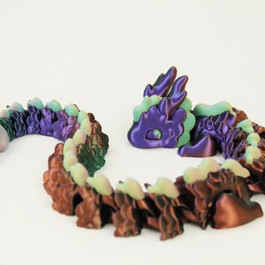 May include: A 3D-printed articulated dragon toy in shades of purple, brown, and green. The dragon is segmented, allowing for flexible posing. The head and tail are separate from the body, and the dragon is set against a white background.