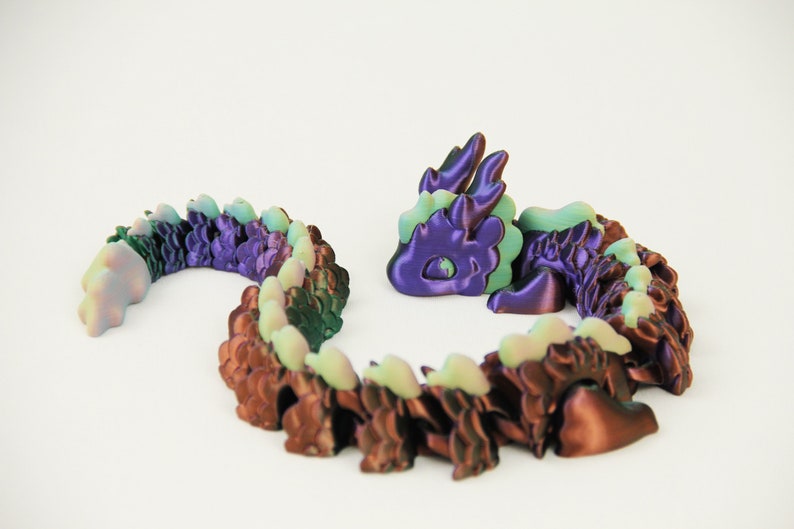 May include: A 3D-printed articulated dragon toy in shades of purple, brown, and green. The dragon is segmented, allowing for flexible posing. The head and tail are separate from the body, and the dragon is set against a white background.