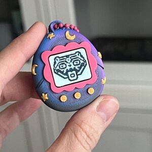 Pode incluir: Um brinquedo digital port&aacute;til para animais de estima&ccedil;&atilde;o em estilo retr&ocirc;, em tons de azul, roxo e rosa. O brinquedo apresenta uma pequena tela exibindo um personagem pixelado dentro de uma moldura rosa. Estrelas e luas douradas decoram o exterior, com um pequeno la&ccedil;o para prender um chaveiro.