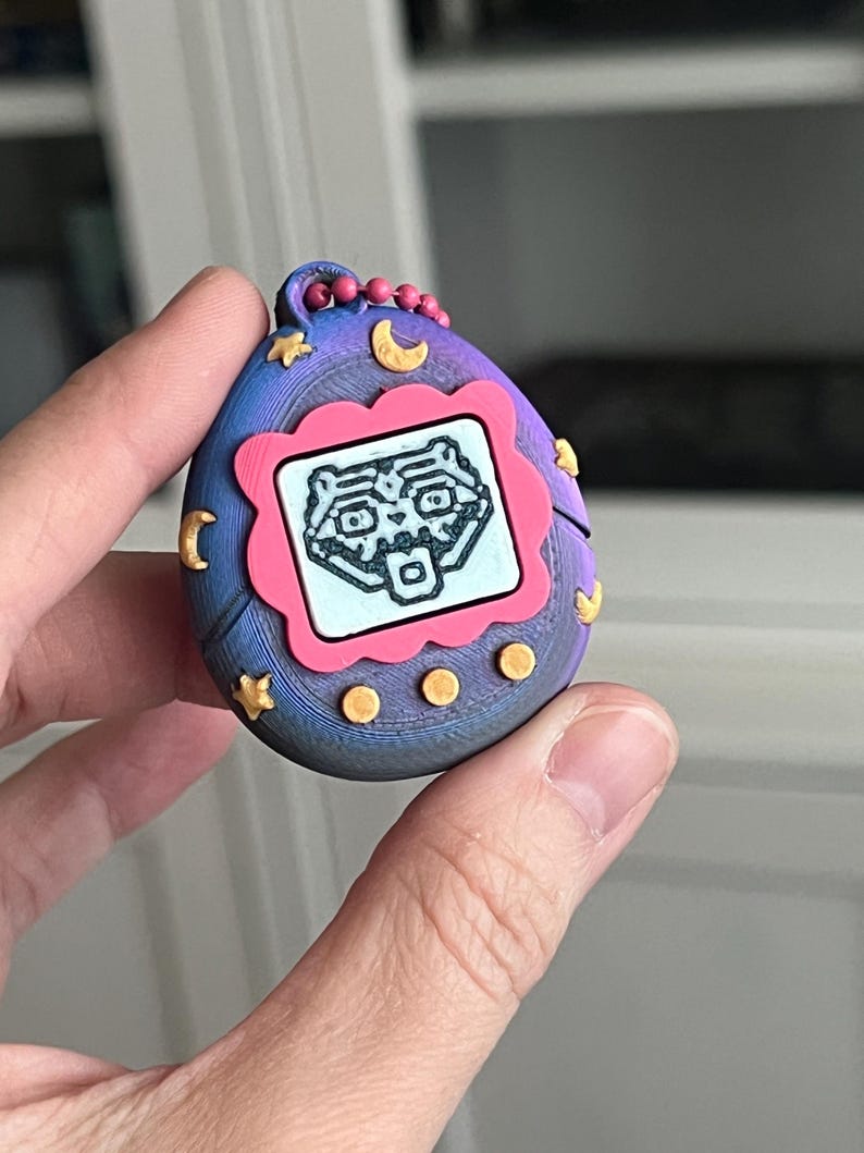 Pode incluir: Um brinquedo digital port&aacute;til para animais de estima&ccedil;&atilde;o em estilo retr&ocirc;, em tons de azul, roxo e rosa. O brinquedo apresenta uma pequena tela exibindo um personagem pixelado dentro de uma moldura rosa. Estrelas e luas douradas decoram o exterior, com um pequeno la&ccedil;o para prender um chaveiro.
