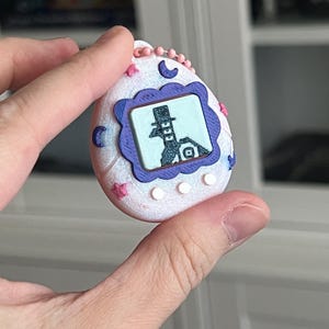 Pode incluir: Um brinquedo digital para animais de estima&ccedil;&atilde;o branco e rosa com uma tela roxa e azul exibindo uma imagem de desenho animado. O brinquedo tem estrelas rosas, luas e uma corrente de contas rosa.