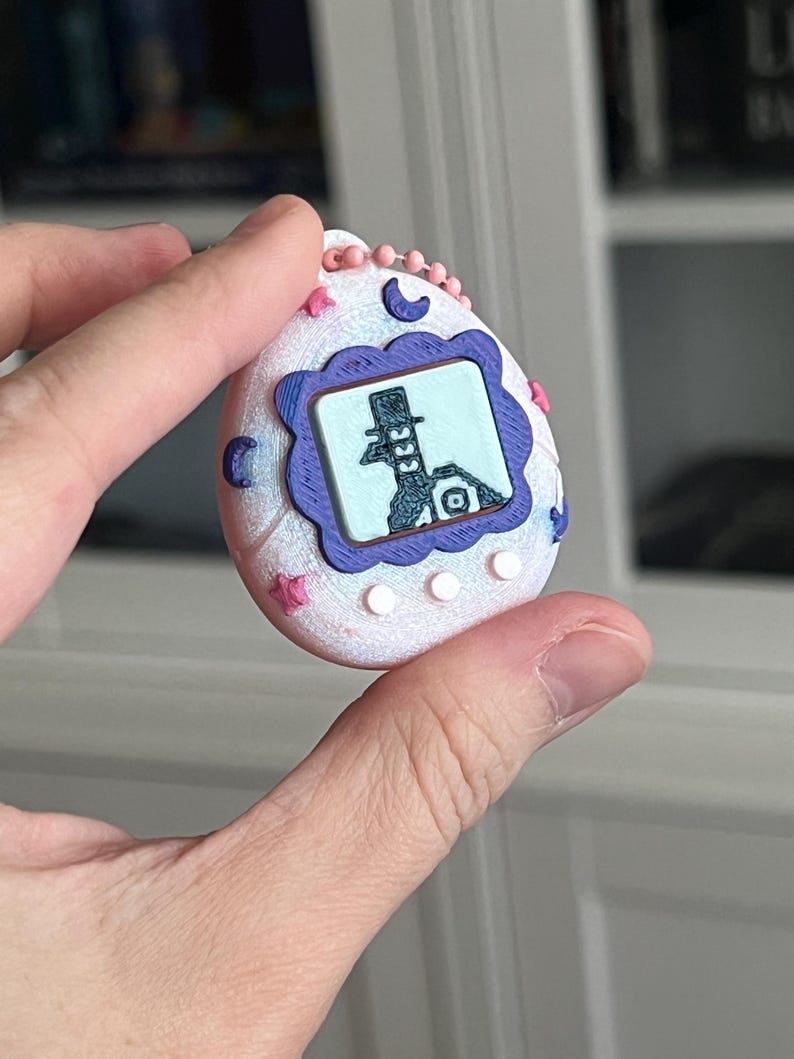 Pode incluir: Um brinquedo digital para animais de estima&ccedil;&atilde;o branco e rosa com uma tela roxa e azul exibindo uma imagem de desenho animado. O brinquedo tem estrelas rosas, luas e uma corrente de contas rosa.