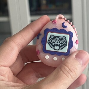 Pode incluir: Um dispositivo digital port&aacute;til rosa com uma tela azul e branca exibindo um rosto de animal de desenho animado. O dispositivo tem uma corrente rosa e &eacute; decorado com estrelas rosas e luas crescentes azuis.