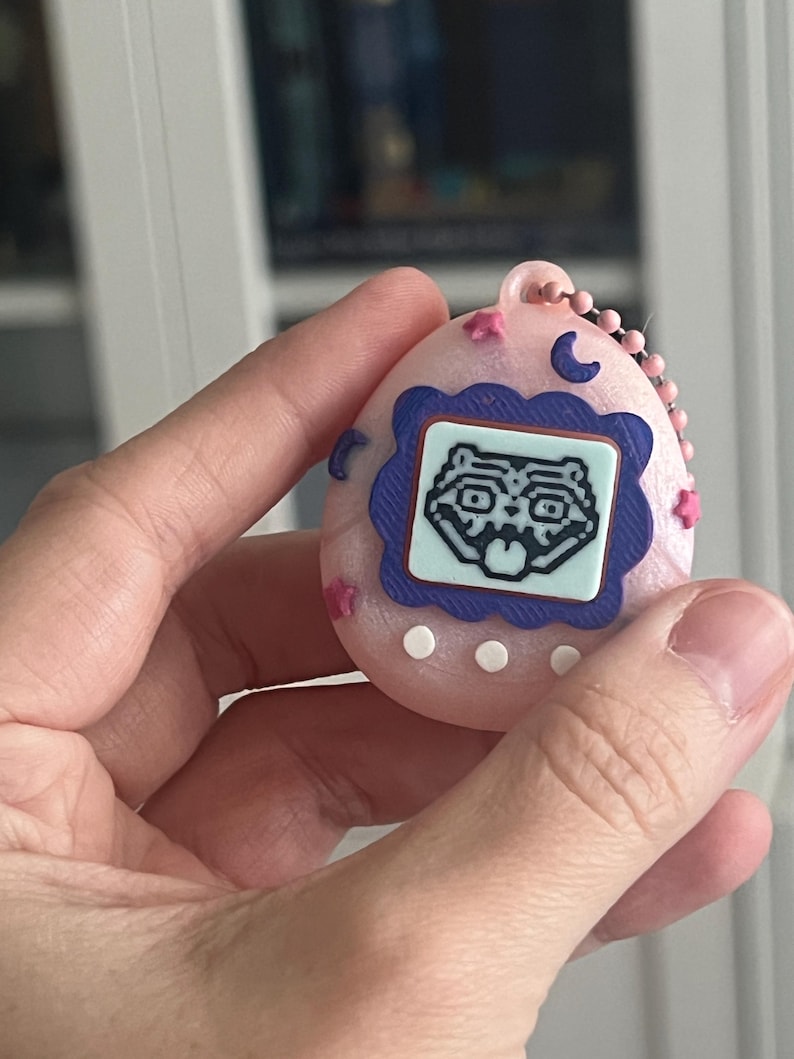 Pode incluir: Um dispositivo digital port&aacute;til rosa com uma tela azul e branca exibindo um rosto de animal de desenho animado. O dispositivo tem uma corrente rosa e &eacute; decorado com estrelas rosas e luas crescentes azuis.