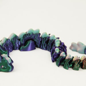 May include: A segmented dragon toy in shades of green, purple, and brown. The articulated design allows for flexible posing. The dragon is made of plastic and has a smooth, glossy finish. The toy is on a white background.