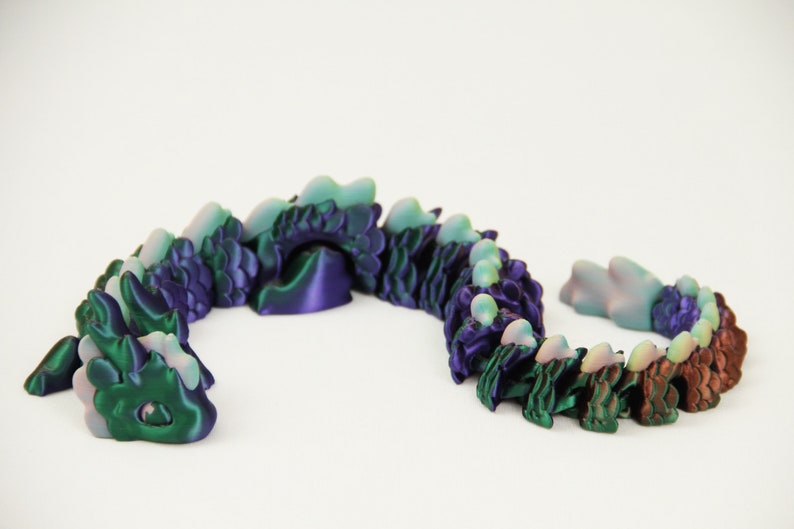 May include: A segmented dragon toy in shades of green, purple, and brown. The articulated design allows for flexible posing. The dragon is made of plastic and has a smooth, glossy finish. The toy is on a white background.