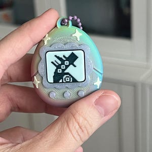 Pode incluir: Um brinquedo digital port&aacute;til em forma de ovo com um gradiente de arco-&iacute;ris pastel. A tela mostra um personagem pixelado dentro de uma moldura decorativa. O brinquedo tem detalhes em forma de estrela e uma corrente roxa para fixa&ccedil;&atilde;o.