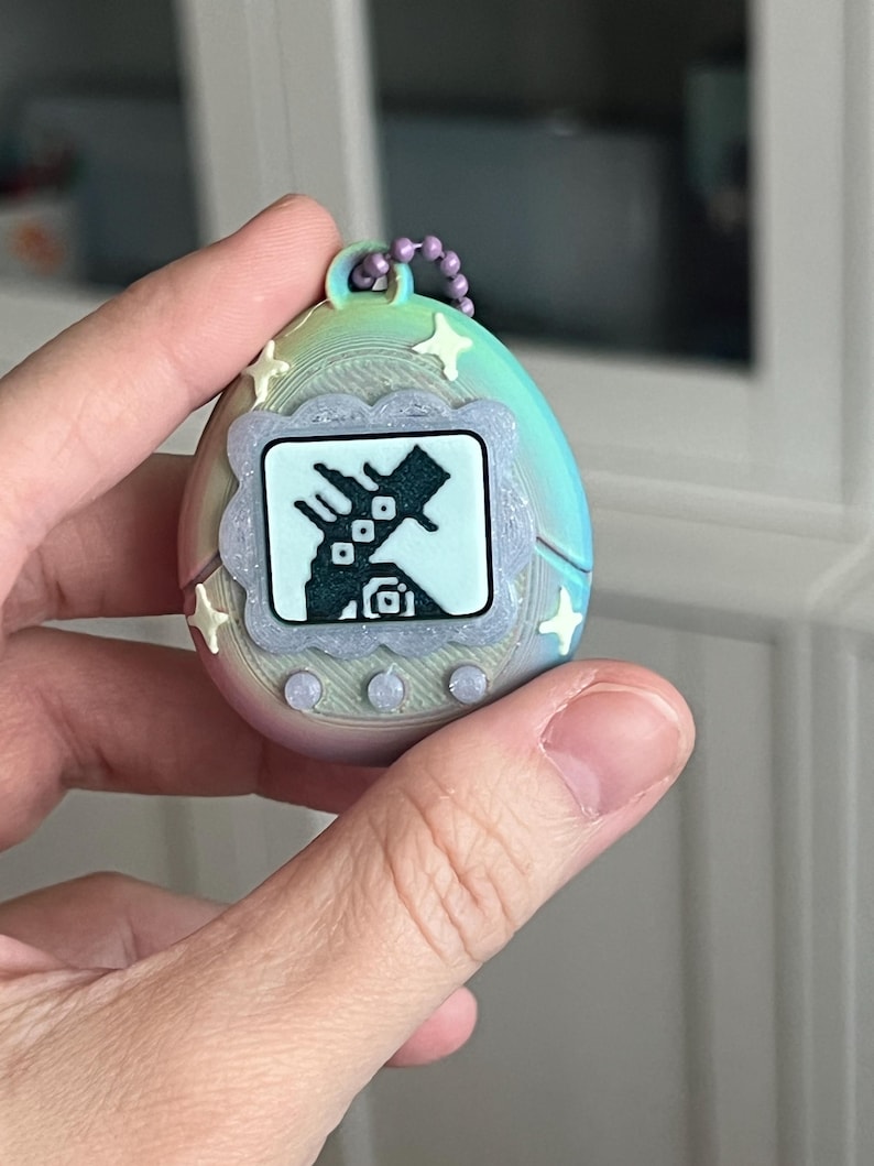 Pode incluir: Um brinquedo digital port&aacute;til em forma de ovo com um gradiente de arco-&iacute;ris pastel. A tela mostra um personagem pixelado dentro de uma moldura decorativa. O brinquedo tem detalhes em forma de estrela e uma corrente roxa para fixa&ccedil;&atilde;o.