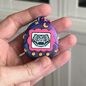 Pode incluir: Um brinquedo digital port&aacute;til roxo e azul, com uma moldura rosa na tela. A tela mostra uma criatura pixelada. O brinquedo &eacute; decorado com luas e estrelas douradas.