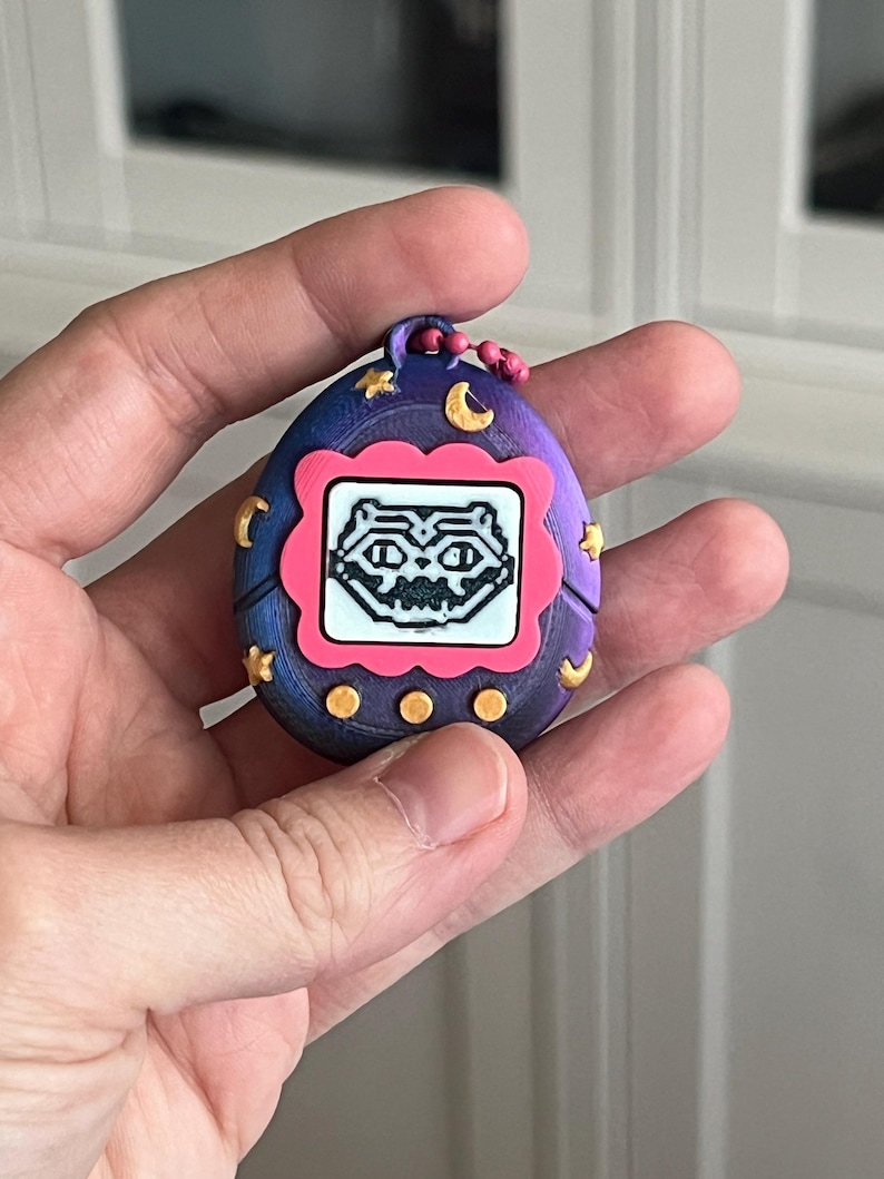 Pode incluir: Um brinquedo digital port&aacute;til roxo e azul, com uma moldura rosa na tela. A tela mostra uma criatura pixelada. O brinquedo &eacute; decorado com luas e estrelas douradas.