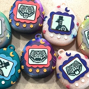 Pode incluir: Uma cole&ccedil;&atilde;o de brinquedos digitais em forma de ovo para animais de estima&ccedil;&atilde;o. Cada brinquedo apresenta um pequeno ecr&atilde; que exibe um personagem pixelizado, emoldurado por uma borda decorativa. Os brinquedos t&ecirc;m uma argola na parte superior para fixa&ccedil;&atilde;o e s&atilde;o decorados com estrelas e luas.