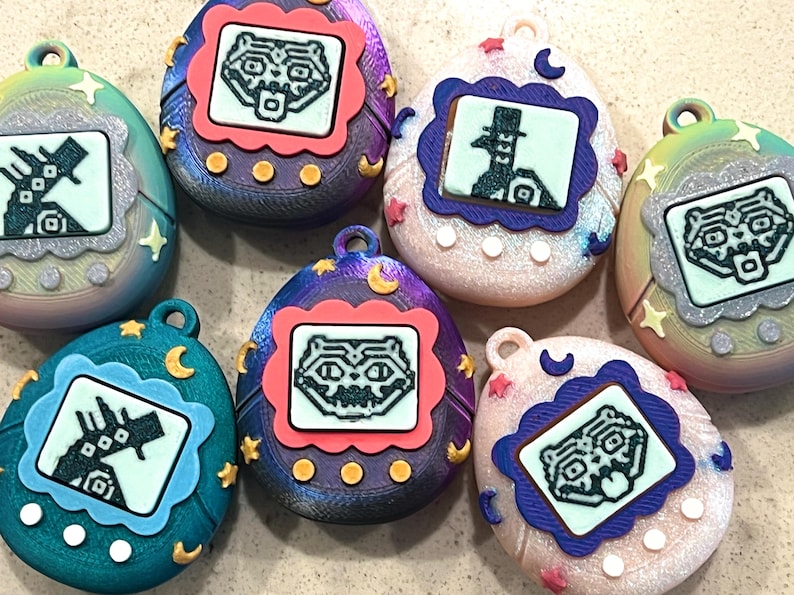 Pode incluir: Uma cole&ccedil;&atilde;o de brinquedos digitais em forma de ovo para animais de estima&ccedil;&atilde;o. Cada brinquedo apresenta um pequeno ecr&atilde; que exibe um personagem pixelizado, emoldurado por uma borda decorativa. Os brinquedos t&ecirc;m uma argola na parte superior para fixa&ccedil;&atilde;o e s&atilde;o decorados com estrelas e luas.