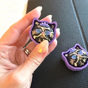 May include: Two small, purple, cat-shaped frames containing a sculpted cat face with black, white, and tan fur patterns. The cat features a small purple crown on its head and blue-tinted sunglasses. The frames are made of a solid purple material.